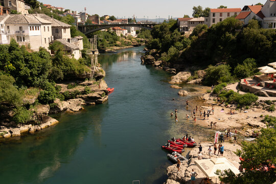 Mostar, Bosnia and Herzegovina - 22 June 2025: View of the Neretva River flowing through the historic city of Mostar, with people swimming and relaxing on the rocky banks near the bridge.