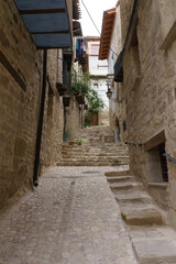 Obraz premium Narrow stone alleyway in historic village. Cobblestone path leads uphill through old buildings. Wooden balconies and hanging plants line the walls. Sunlight filters through arches above