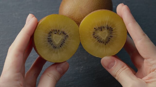 Macro shot of female hands holding two halves of ripe golden kiwi, gently pressing and rotating them together. Rich juicy pulp with playful movement for healthy eating, gourmet food, culinary content.