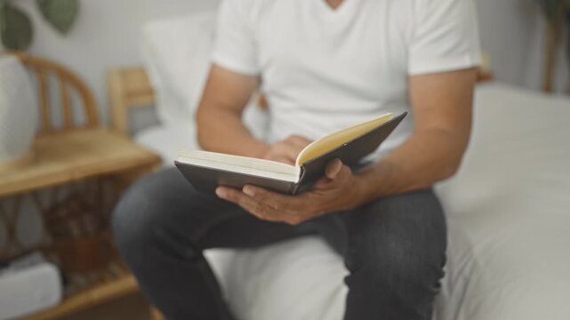 Man in white t shirt holding open book with visible hands turning page while sitting on bed in bedroom; quiet reflection calm.