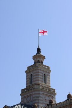 Georgian national flag flying above a historic ornate tower in Tbilisi, Georgia, against a clear blue summer sky.