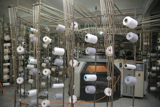 Lahore, Pakistan - 02 October 2024: View of the Quaid-e-Azam Industrial Estate textile factory, featuring industrial knitting machines surrounded by numerous spools of white and grey yarn.