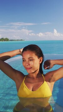 Luxury swimming pool Asian bikini model woman wearing yellow fashion swimsuit relaxing in infinity pool at overwater bungalow resort, tropical vacation. Vertical video of real woman without makeup