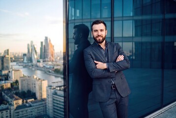 Fototapeta premium Confident Young Businessman in Elegant Suit Standing on High Rise Balcony with Modern Cityscape View