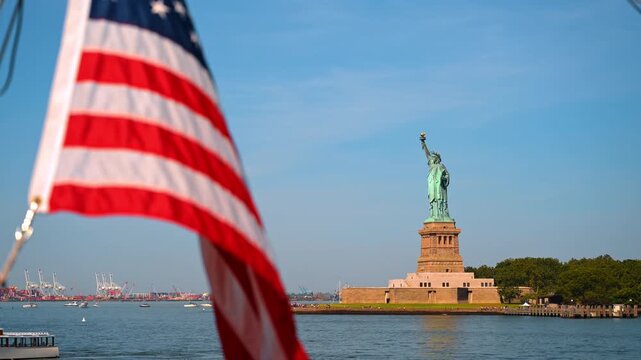 New York, USA, 4 Jule 2026 : American flag framing Statue of Liberty. Shipping cranes line the distant shore as boats cross the harbor under a clear blue sky.