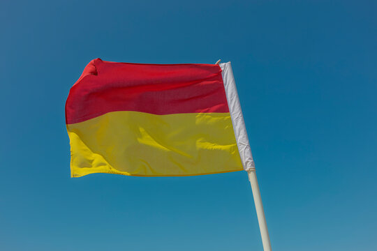 Red and yellow beach safety flag stretched in the wind above a Cornish beach under an empty blue sky.