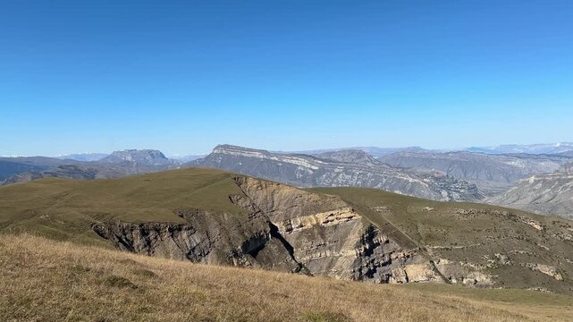 High-elevation panoramic scene showing dry grassy slope and low rolling ridge in foreground that drops to exposed sedimentary outcrop and steep cliff faces with visible horizontal bedding