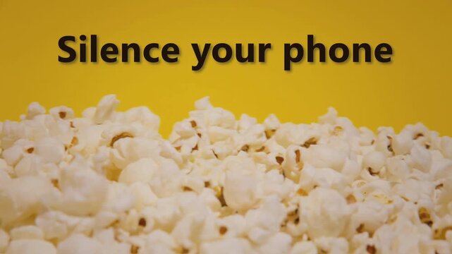 Plain popcorn fills frame on bright yellow surface with silence your phone text overlay. Movie theater preshow snack announcement with simple clean design for cinema etiquette.