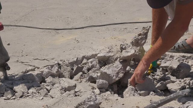A construction worker uses pliers to cut exposed steel reinforcement wire and separate broken concrete while wearing one glove, shorts and open-toe sandals, illustrating unsafe demolition practices.