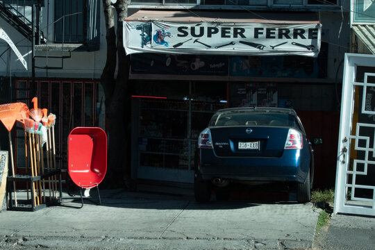 Mexico City, Mexico - January 27, 2024 - A parked Nissan Sentra sits outside "S&uacute;per Ferre," a hardware store, with garden tools and a wheelbarrow displayed on the sidewalk in Mexico