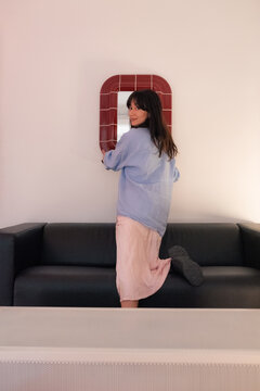 Woman hanging mirror on wall and looking at camera