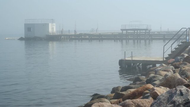 Wooden pier with swim ladder stands on rocky shore in dense coastal morning fog. White bathhouse pavilion sits in calm sea water. This quiet seaside spot offers outdoor wellness and cold plunge.