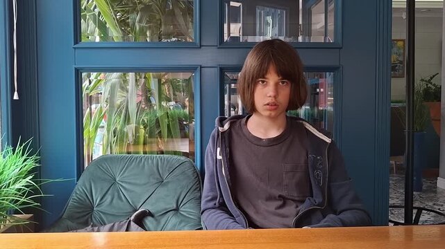 13 year old boy sits alone in a cafe with plants and blue walls around him