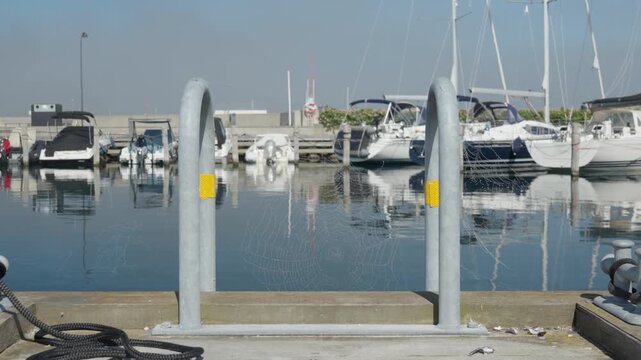 Morning dew drops glisten on a spider web across the dock ladder. Moored yachts float in the calm marina harbor during sunny dawn. Quiet seaside scenery reflects tranquil coastal freshness.
