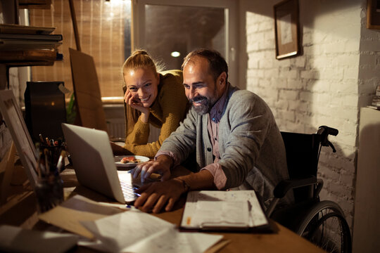 Smiling couple with man in wheelchair working on laptop at home office at night