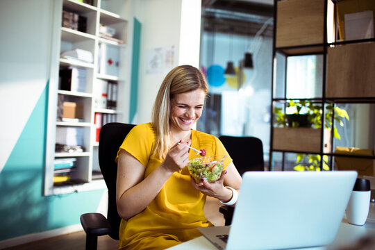 Smiling woman eating salad at office desk with laptop