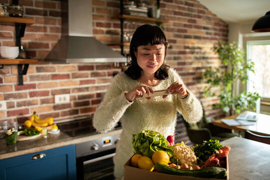 Woman photographing fresh grocery delivery in home kitchen