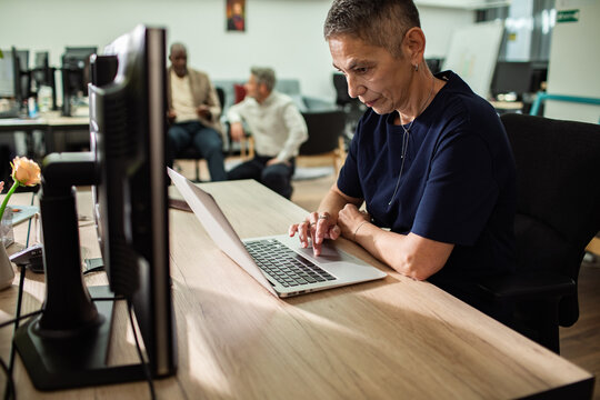 Mature businesswoman working on laptop in modern open office with colleagues in background