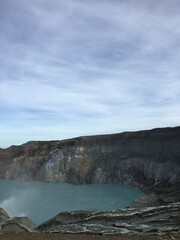 Volcanic crater lake with turquoise water and rocky cliffs under a cloudy sky © Design_Vr