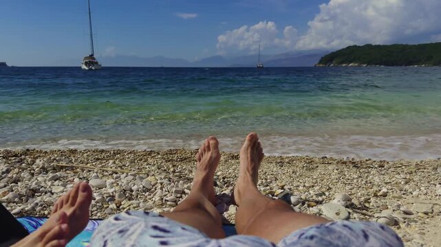 Person Feet Relaxing On Pebble Beach Facing Clear Turquoise Sea Water Against Sailboat Yachts. Relaxation And Travel 