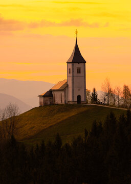 Sts. Primus and Felician Catholic Church near Jamnik, Slovenia, on a hilltop at sunset, surrounded by autumn forest, rolling hills and dramatic mountain silhouettes.