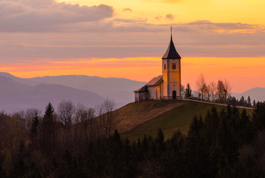 Sts. Primus and Felician Catholic Church near Jamnik, Slovenia, on a hilltop at sunset, surrounded by autumn forest, rolling hills and dramatic mountain silhouettes.