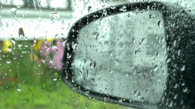 Car side view mirror covered in rain droplets reflecting green grass and a pink object on a wet rainy day.