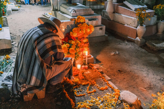 Santa Cruz Xoxocotlan, Oaxaca, Mexico - 31 October 2023: View of a man in a traditional poncho kneeling at a grave decorated with marigold flowers and candles during Dia de los Muertos.