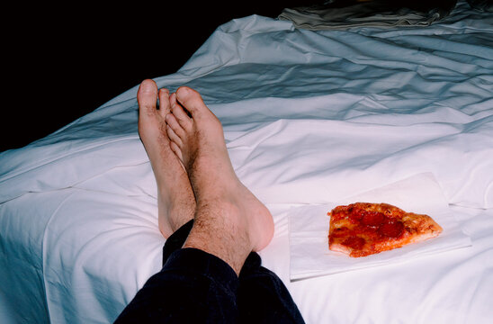 a man relaxes on a bed beside a slice of pepperoni pizza