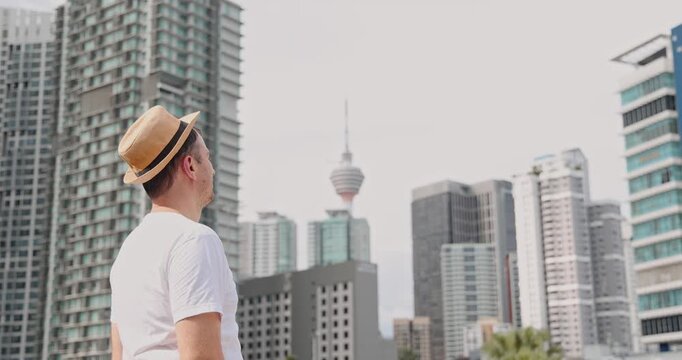 Man in straw hat and white t-shirt observing Kuala Lumpur city skyline with KL Tower, urban tourism and travel exploration in Malaysia, commercial lifestyle .