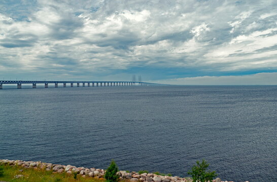Sibbarp, Sweden - 30.07.2017: Long &Oslash;resund Bridge spanning deep blue waters under a heavy overcast cloudy sky with rocky shoreline foreground