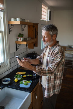 Middle-aged man using smartphone while prepping healthy breakfast at home kitchen