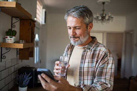 Middle-aged man drinking water while using smartphone in home kitchen