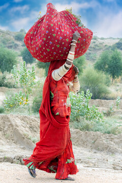 Nagarparkar, Pakistan - 23 December 2019: View of a woman in a traditional red dress carrying a large bundle on her head while walking through the rural landscape of Nagarparkar.