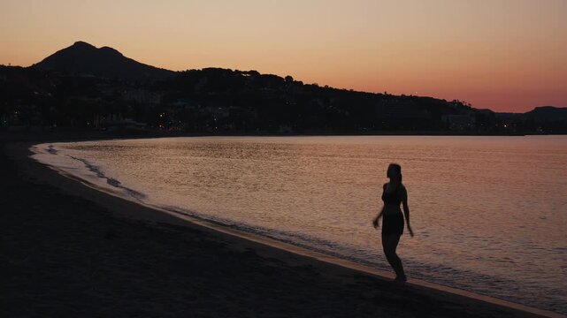 Energetic woman performing jumping jacks at dusk on a beach, a dynamic silhouette against the calm ocean and distant town lights, inspiring fitness and active lifestyle.