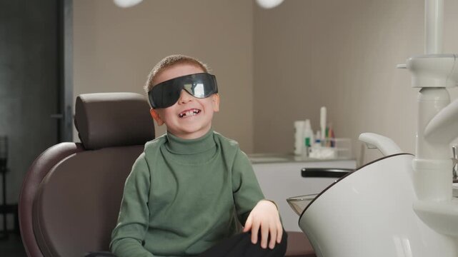 Young boy sits in dental chair putting on protective eyewear. Dental equipment and supplies are visible around him as boy prepares for orthodontic procedure in clinic setting