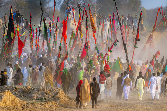 Okara, Punjab, Pakistan - 13 March 2024: View of the annual Urs and mela at the shrine of Hazrat Daud Bandagi Kirmani, with a crowd of devotees carrying colorful flags in a dusty setting.
