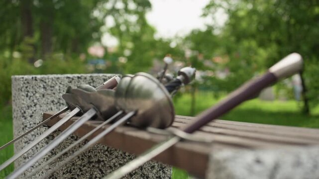 Close up of several fencing swords resting on wooden bench in outdoor park setting. gloved hand briefly touches one sword emphasizing metallic details and craftsmanship against green foliage