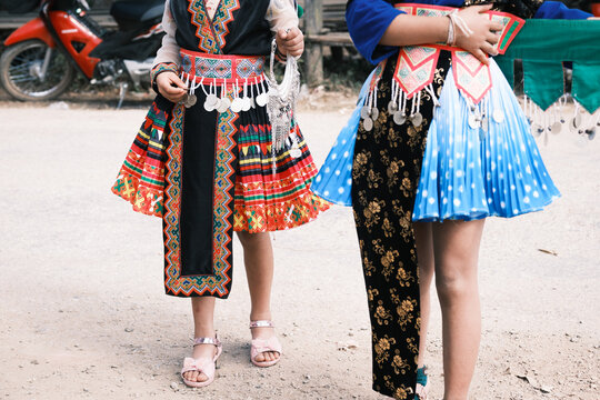 Laos - 30 December 2024: View of two people in traditional Hmong tribe clothing with intricate embroidery, colorful pleated skirts, and silver coins, standing near a red motorcycle.