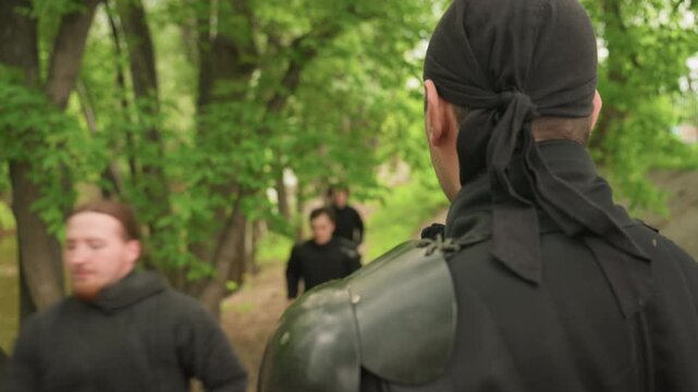 Multiple men in dark clothing run along forest path observed by another men wearing armor. scene features dense green foliage and trees lining dirt trail with soft natural light filtering