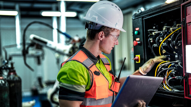 A robotics engineer uses a laptop to run diagnostics on an AI system. He is checking the hardware, wiring, and high-tech sensors of an industrial robot.