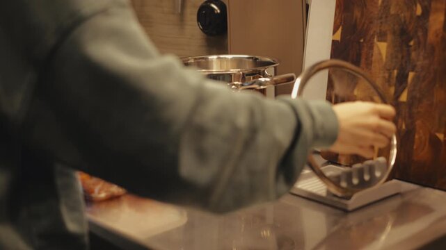 Close-up of woman hands placing empty stainless steel pot on portable induction stove and moving glass lid onto pot lid stand, preparing to cook for home food routine in bright modern loft kitchen.