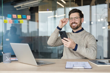 Obraz premium Confident businessman adjusting his spectacles while smiling at the camera, holding a smartphone and sitting at his desk with a laptop in a contemporary corporate workspace