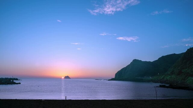 A beautiful sunrise over the calm sea at Dawulun Beach in Keelung, Taiwan, with a small island on the horizon and green hills.