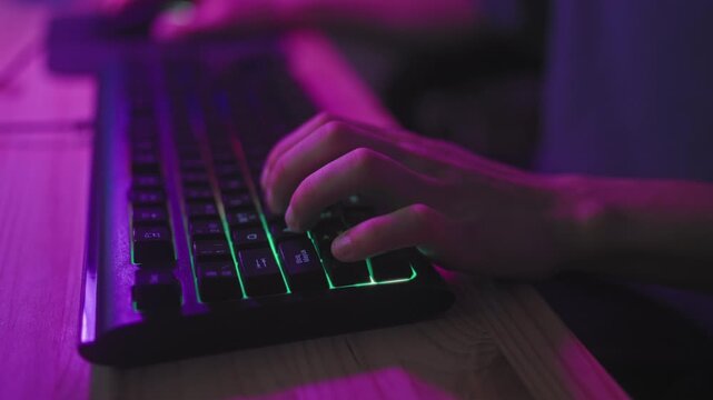 Young hispanic man typing with hands on rgb backlit gaming keyboard in studio at wooden desk under purple neon lighting; focused immersion.