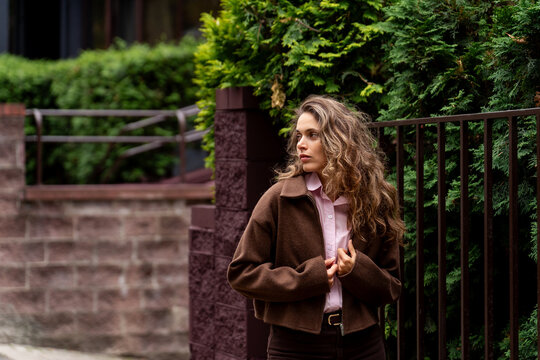 Young woman in casual brown jacket standing by green hedge