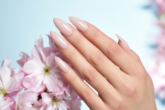Elegant feminine hand with almond-shaped nails posed beside delicate pink cherry blossoms against a soft blue background, showcasing a flawless blush-pink manicure