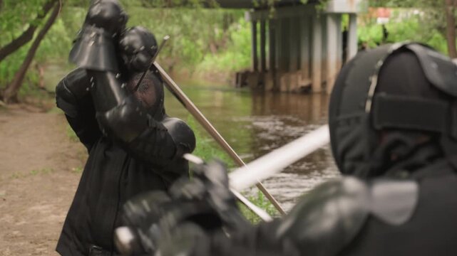 Two people in full black fencing gear engage with swords by riverbank under overpass demonstrating controlled movements and techniques. Their protective mesh masks cover faces
