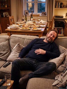 A relaxed man reclines on a cozy sofa after a large dinner, holding his stomach while a table full of leftover food and drinks stands in the background.