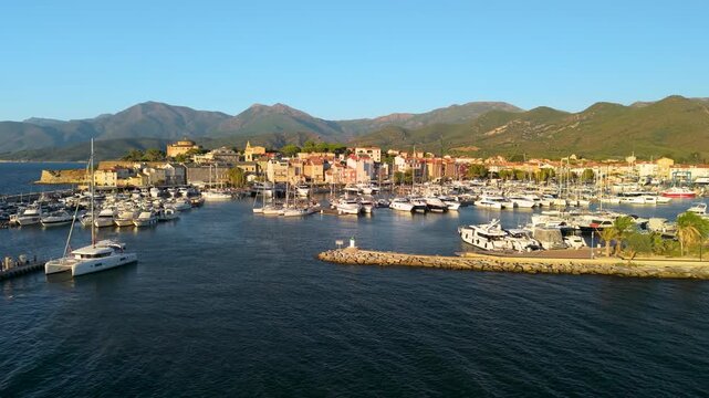 Saint Florent Corsica 4K drone video part 1. Sunset panning view to moored yachts in harbor. Cinematic fly in toward coastal marina and sailboats in golden hour light. Travel b roll stock footage